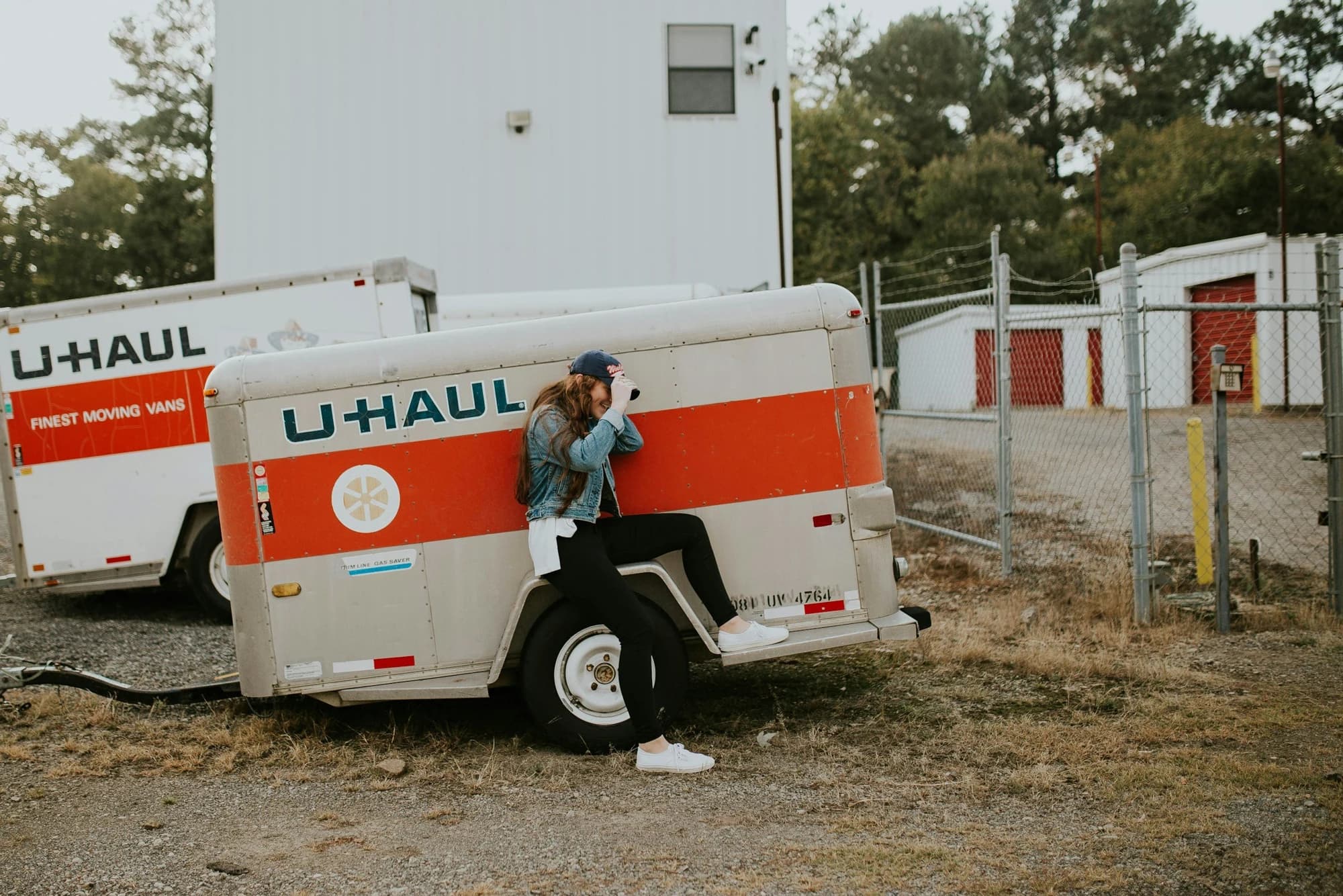 A person beside a U-Haul trailer, with a moving truck and storage units behind